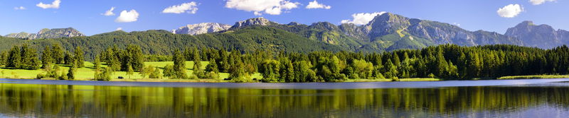 Bergsee vor bewaldeten Ufern und einer Gebirgskette unter blauem Himmel