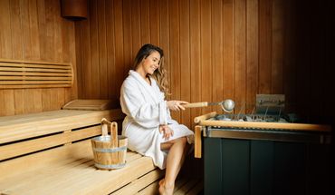 Frau im Bademantel sitzt auf der Holzbank einer Sauna neben Aufgusskübel und Kelle