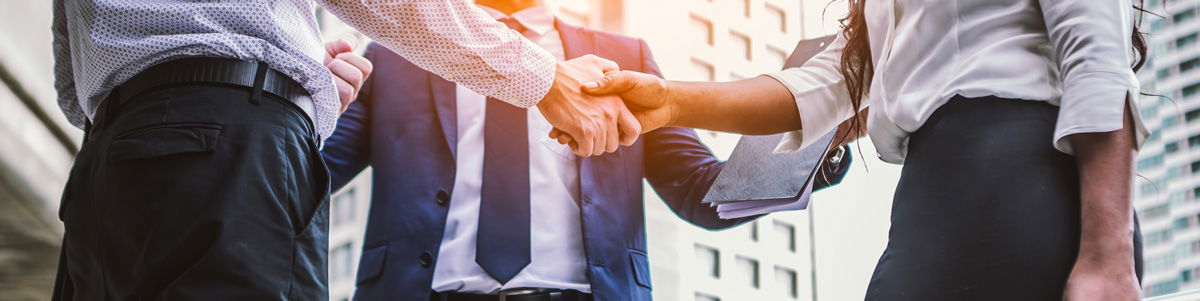 Drei Geschäftspersonen in formeller Kleidung geben sich vor modernen Bürogebäuden die Hand und symbolisieren eine geschäftliche Zusammenarbeit oder einen Vertragsabschluss.
