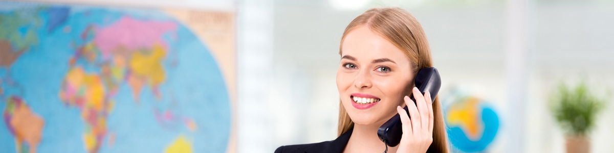 Eine lächelnde Mitarbeiterin in Businesskleidung telefoniert in einem Büro; im Hintergrund sind eine Weltkarte und ein Globus zu sehen.
