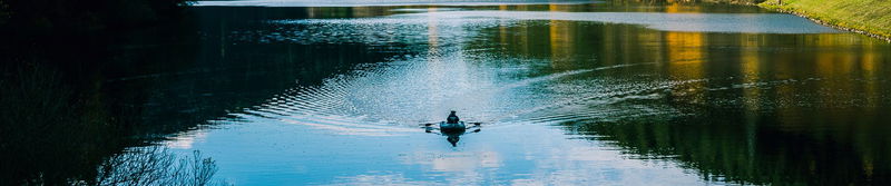 Person allein im Ruderboot auf einem stillen See mit Spiegelungen der Uferbäume